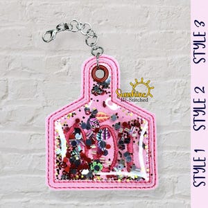 May include: A pink, glitter-filled keychain in the shape of a cow tag. The tag has a clear center with colorful confetti and the word "Mom" visible. A silver chain and clasp are attached to the top. The image also shows three tag style options.
