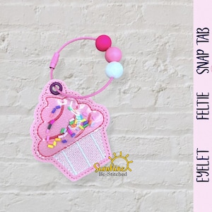 Op de afbeelding: Roze cupcake-vormige feltie met kleurrijke sprinkles en een roze kralenband. De feltie heeft een helder venster en is bevestigd aan een roze koord met roze, witte en fuchsia kralen. De afbeelding toont ook cupcake-illustraties.