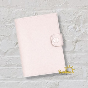 May include: A light pink, rectangular journal or planner with a button closure. The cover is smooth and has a stitched border. The journal is closed with a matching pink button. The logo "Sunshine Be-Stitched" is in the bottom right corner.