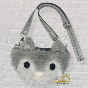 May include: A grey crossbody bag in the shape of a wolf's face, with fluffy ears and a stitched face. The bag has a grey strap with decorative stitching and a metal buckle. The bag is on a white brick wall.