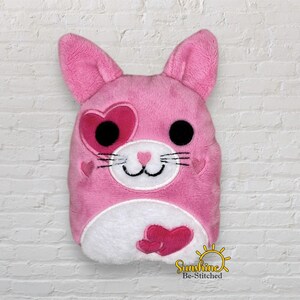 May include: A pink plush bunny with heart-shaped accents. The bunny has large black eyes, a white muzzle, and a heart-shaped eye. A heart is on the cheek and on the belly. The bunny has long ears and a cute design.
