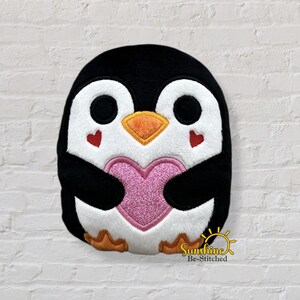 Può includere: Un pinguino di peluche con corpo nero, faccia bianca e becco arancione. Il pinguino tiene in mano un cuore rosa e ha accenti a forma di cuore rosso. Il design è ricamato e ha un aspetto morbido e strutturato.