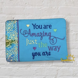 May include: A rectangular, light blue decorative item with the embroidered text "You are Amazing just the way you are." The item has a floral design on the left side and small heart accents. The logo "Sunshine Be-Stitched" is in the bottom right corner.
