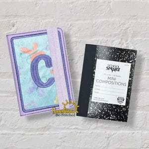 May include: An embroidered notebook cover with the letter "C" and a composition notebook. The notebook cover is light blue with floral and butterfly designs, a purple border, and a purple elastic band. The composition notebook is black and white.