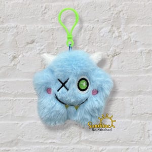 May include: A light blue plush monster keychain with a green clip. The monster has white horns, a winking eye, and a smiling mouth. Pink blush dots are on the cheeks. The text "Sunshine Be-Stitched" is at the bottom.