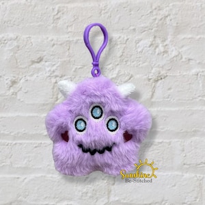May include: A plush, lavender monster-shaped keychain with three blue eyes, white horns, and a black stitched mouth. The keychain has a purple clip at the top. The design includes small red heart details on the cheeks. The logo "Sunshine Be-Stitched" is visible.