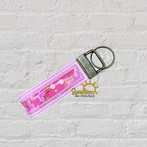 May include: A pink fabric keychain featuring a strawberry pattern and a clear plastic window, accented with white circular embellishments. The keychain is attached to a dark bronze-toned metal hardware ring and includes the text Sunshine Be-Stitched.