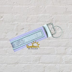 Può includere: Un portachiavi rettangolare con un bordo in tessuto viola chiaro che circonda una striscia centrale di materiale argentato glitterato. Il portachiavi è fissato a una chiusura in metallo color argento e a un anello circolare. Il testo Sunshine Be-Stitched è visibile.