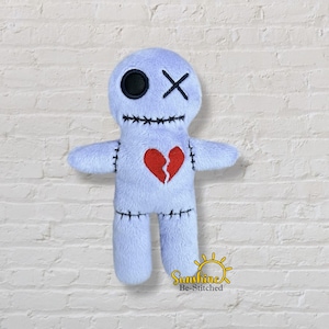 May include: A pale blue plush voodoo doll featuring a red broken heart, black stitching, and a black eye. The doll has an X for a mouth and an X for an eye. It is set against a white brick wall.
