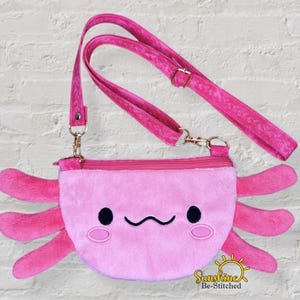May include: A pink axolotl-shaped crossbody bag with a zippered top and a long, adjustable strap. The bag features a smiling face with black eyes and a curved mouth, and pink appendages. The bag is made of soft, plush material.