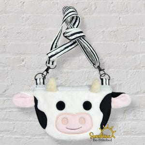 May include: A white and black cow-shaped crossbody bag with a striped strap. The bag features a cow face design with black spots, pink ears, and a pink nose. The strap is black and white striped.