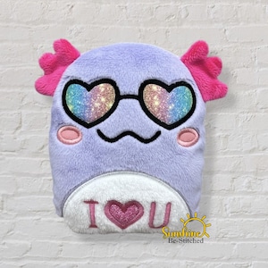 May include: A plush, lavender-colored creature with pink accents and heart-shaped sunglasses. The creature has a white belly with the text "I ❤️ U" embroidered in pink. The image is against a white brick wall.