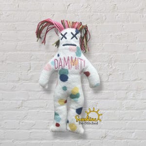 May include: A white plush doll with multicoloured polka dots, featuring embroidered X-shaped eyes and a straight line mouth. The word DAMMIT! is embroidered in pink on the chest. The doll has yarn hair in shades of pink, green, and brown.