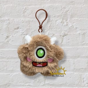 May include: A brown, plush monster keychain with a single green eye, white horns, and a smiling red mouth. The keychain has a brown loop and a gold-colored clasp. The text "Sunshine Be-Stitched" is in the lower right corner.