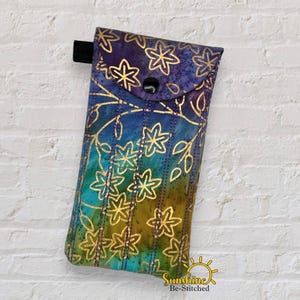 May include: A fabric glasses case with a floral design. The case has a flap with a black button closure and a black loop on the back. The case has a gradient of colours, including blue, green, and gold.