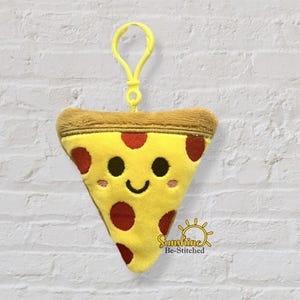 Op de afbeelding: Een pluche pizzasnede sleutelhanger met een lachend gezicht, rode pepperoni en een bruine korst. De gele sleutelhanger heeft een gele clip aan de bovenkant voor eenvoudige bevestiging. Het item staat tegen een witte bakstenen muur.