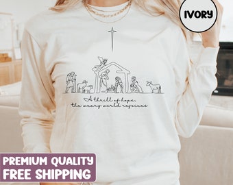 May include: A white long-sleeved sweatshirt with a black line drawing of the nativity scene. The text "A thrill of hope, the weary world rejoices" is printed below the image. The sweatshirt is available in ivory.