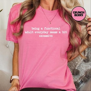 May include: A bright pink t-shirt with the text "being a functional adult everyday seems a bit excessive" in white. The shirt has short sleeves and a crew neck. A small, round logo with the words "Crunch Berry" is in the upper right corner.
