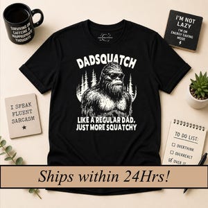 Può includere: T-shirt nero con la scritta "Dadsquatch" e una grafica di un Bigfoot con occhiali da sole. La maglietta dice anche "Like a regular dad, just more squatchy." Altri oggetti includono una tazza, un cartello con "I speak fluent sarcasm" e un blocco note.