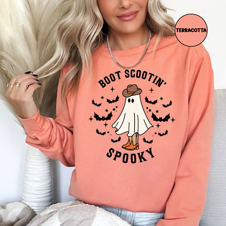 May include: A terracotta orange long-sleeved sweatshirt with the text "Boot Scootin' Spooky" and a ghost wearing a cowboy hat and boots. The ghost is surrounded by bats and stars.