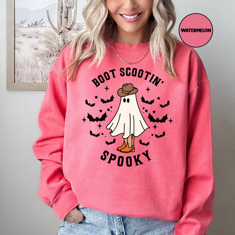 May include: A pink sweatshirt featuring a ghost wearing a cowboy hat and boots. The text on the shirt reads "Boot Scootin' Spooky".