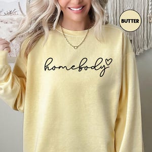 May include: A yellow sweatshirt with the text "homebody" in black script with a small black heart to the right of the word.