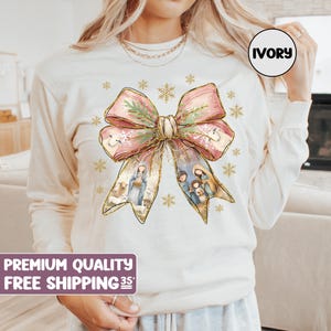 Puede incluir: Una sudadera blanca de manga larga con un diseño de lazo rosa y dorado. El lazo tiene una escena de la natividad en su interior. El texto "IVORY" está en un cuadro blanco en el lado derecho de la imagen.