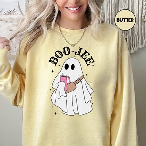 May include: A yellow sweatshirt with the text "BOO-JEE" in black letters. A white ghost with a pink drink and a brown bag slung over its shoulder is in the centre of the sweatshirt.