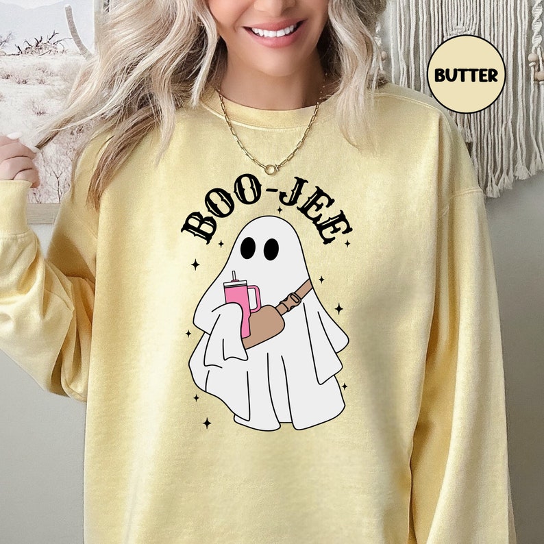 May include: A yellow sweatshirt with the text "BOO-JEE" in black letters. A white ghost with a pink drink and a brown bag slung over its shoulder is in the centre of the sweatshirt.