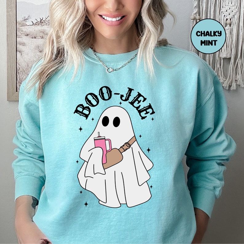 May include: A light blue sweatshirt featuring a white ghost holding a pink drink and a brown bag slung over its shoulder. The text "BOO-JEE" is written in black above the ghost.