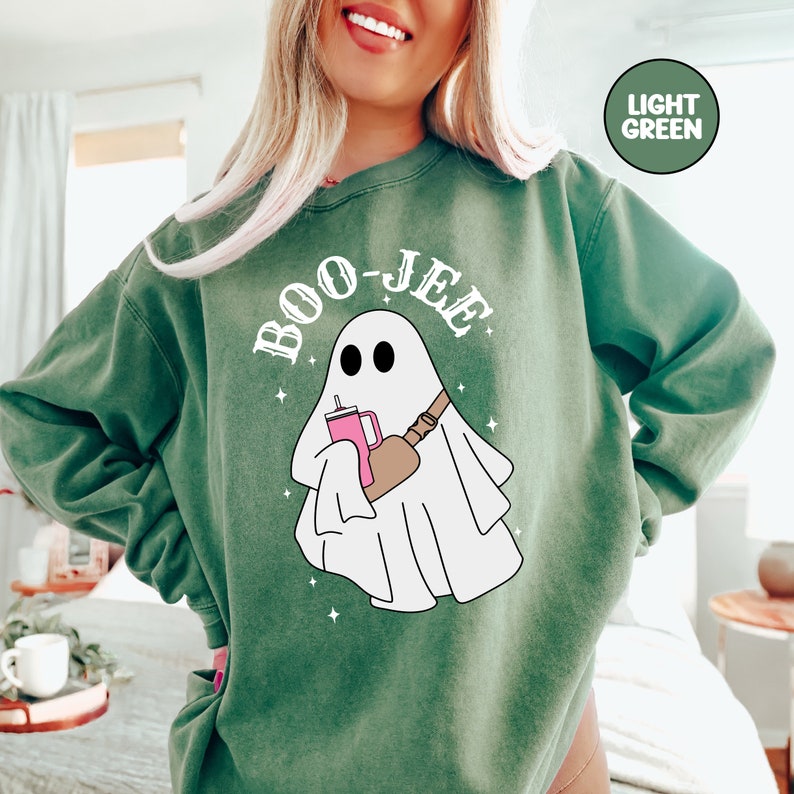 May include: A light green sweatshirt featuring a white ghost holding a pink drink and a brown bag. The text "BOO-JEE" is written in white above the ghost.