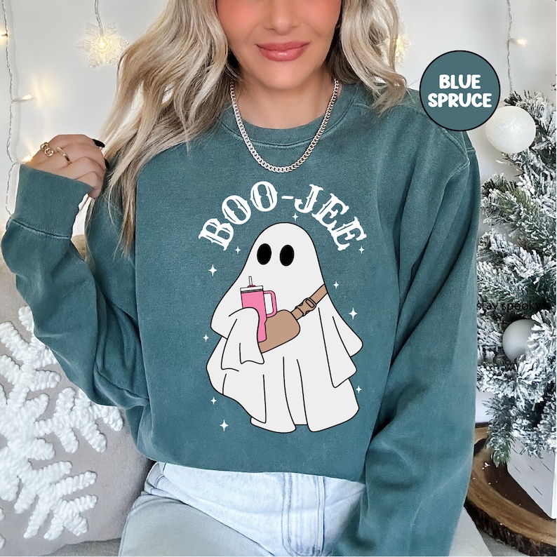 May include: A teal blue sweatshirt with a white ghost holding a pink drink and a brown bag. The text "BOO-JEE" is printed in white on the sweatshirt.