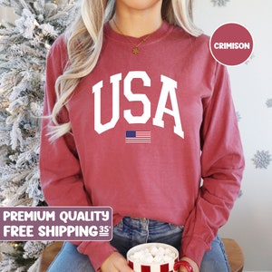 May include: A long-sleeve crimson sweatshirt featuring the white text USA in a collegiate font centered on the chest, positioned above a small graphic of the American flag. The garment has a crew neckline and a solid, muted red fabric finish.