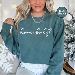 May include: A teal blue sweatshirt with the text "homebody" in white cursive lettering with a small heart symbol. The sweatshirt is a cropped style and has long sleeves.