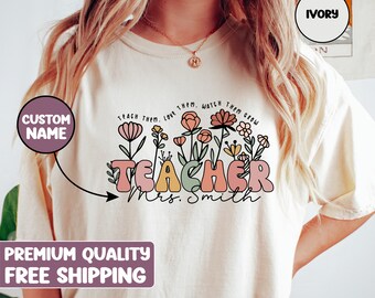 Pode incluir: Camiseta de cor marfim com um design floral e o texto "TEACH THEM. LOVE THEM. WATCH THEM GROW" acima da palavra "TEACHER" em letras coloridas e contornadas. O texto "Mrs. Smith" está abaixo da palavra "TEACHER".