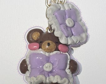 May include: A handmade brown bear charm with a purple bow and matching pillow-shaped accessory. The bear has pink cheeks and a gold-colored chain for hanging. The charm is made of clay and has a whimsical, handcrafted appearance.