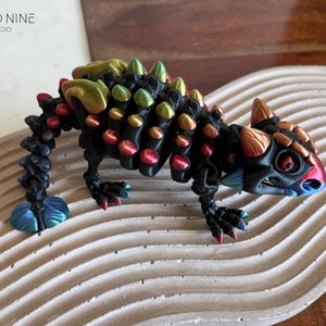 May include: A colorful, stylized lizard figurine with a black body and vibrant accents of red, blue, green, and gold. The figurine is posed on a textured, wavy surface. The text "CLOUD NINE 3D STUDIO" is in the upper left corner.