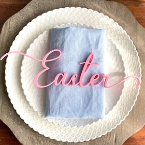 May include: Pink acrylic "Easter" sign on a blue linen napkin, placed on a white plate with a decorative edge.