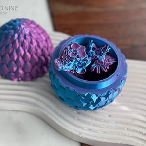 May include: A blue and purple dragon egg-shaped trinket box with a detailed, scaly exterior. The lid is open, revealing a small, purple and blue dragon figurine inside.