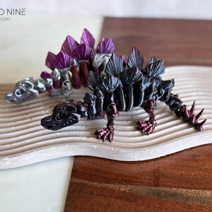 May include: Two 3D-printed Stegosaurus dinosaur models. One is black with red accents, the other is silver and purple. They are displayed on a wavy, light-colored surface with a wooden background. The Cloud Nine 3D Studio logo is in the upper left.