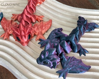 May include: Two small, articulated dragon figurines. One is red with orange accents and the other is black with purple accents. Both have wings and are posed with their heads tilted up.