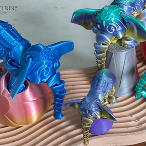 May include: Five 3D printed blue, green, and gold elephant figurines. The elephants are standing on various colorful bases. The elephants have intricate details and are painted in a metallic finish. The elephants are arranged on a textured surface.