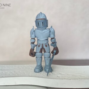 May include: A gray, plastic toy knight figurine wearing armor and holding a sword. The figurine is standing on a white, textured surface.