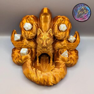 May include: A gold-colored 3D printed dice tower shaped like a nine-tailed fox with a central compartment for dice. The fox has a detailed face and each tail has a compartment for dice.