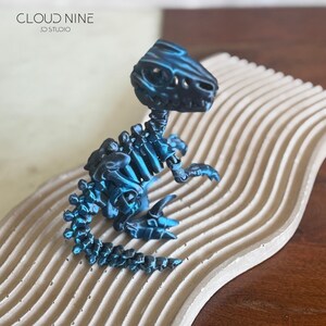 May include: A blue and black 3D-printed dinosaur skeleton figurine. The figurine is posed on a beige, wavy, textured surface. The text "CLOUD NINE 3D STUDIO" is in the upper left corner.
