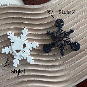 May include: Two styles of snowflake ornaments with a skull design. The first style is white and the second style is black. The ornaments are on a wavy, light brown surface. The text "Style 1" and "Style 2" are written on the surface.