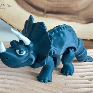 May include: A teal blue 3D printed toy triceratops dinosaur with white horns and eyes. The dinosaur is posed walking with its head turned to the side.