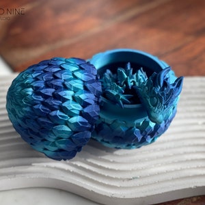 May include: A blue and teal dragon egg-shaped trinket box with a dragon head peeking out of the top. The box is made of a textured material that resembles scales.