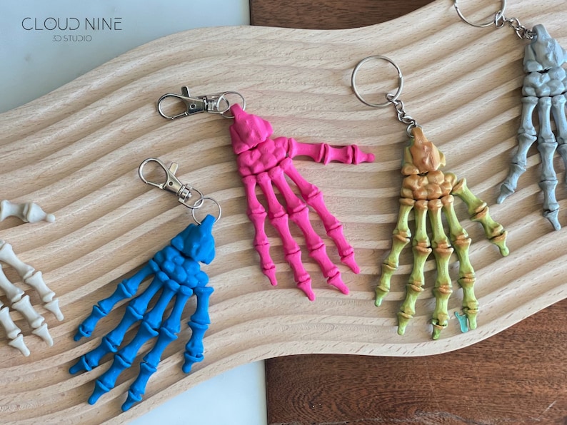 May include: Five skeleton hand keychains in different colors: blue, pink, gold, gray, and white. Each keychain has a silver metal ring and a silver metal clasp.