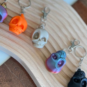 May include: Five colorful skull keychains with silver key rings. The skulls are made of plastic and have different colors and finishes, including orange, gray, black, and iridescent blue and purple.